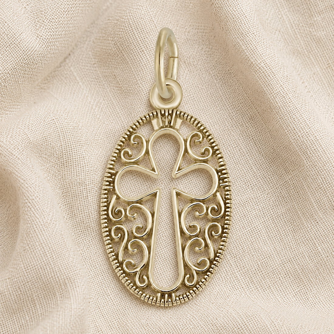 The Filigree Oval Cross Charm in gold plate by Rembrandt Charms, featuring an ornate cross design with intricate scrollwork, is displayed on a beige fabric background.