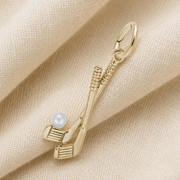 A unique Rembrandt Charms Golf Clubs-Yellow Gold 14k Charm features two crossed golf clubs with a white pearl at the base, displayed on a folded beige fabric background—a distinctive design that makes for a memorable golfer keepsake.