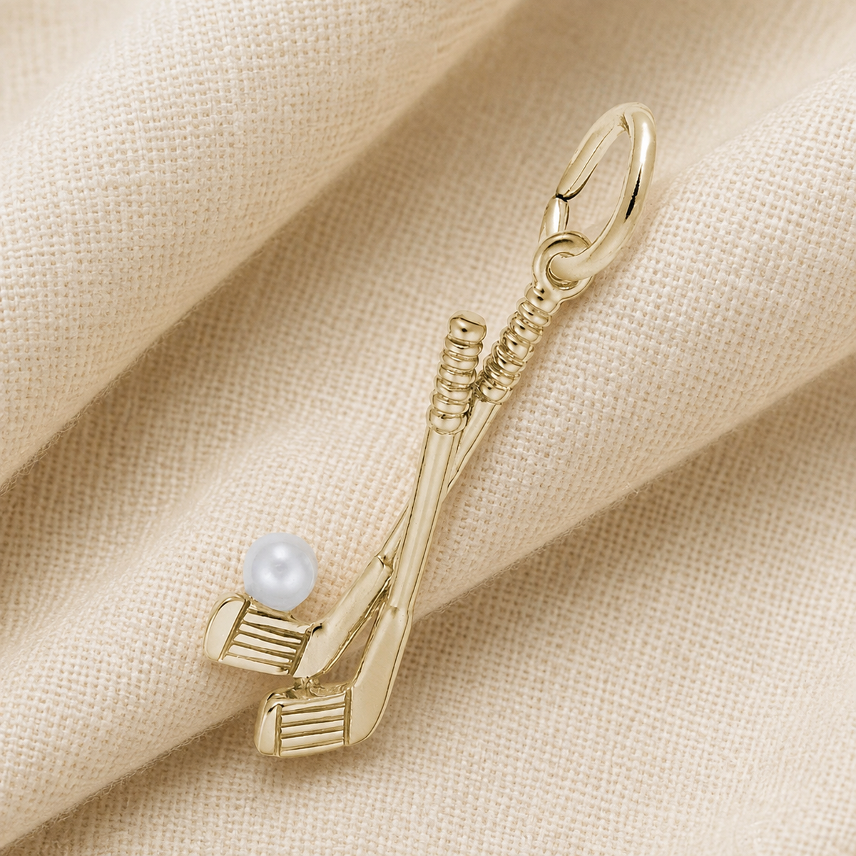 A unique Rembrandt Charms Golf Clubs-Yellow Gold 14k Charm features two crossed golf clubs with a white pearl at the base, displayed on a folded beige fabric background—a distinctive design that makes for a memorable golfer keepsake.
