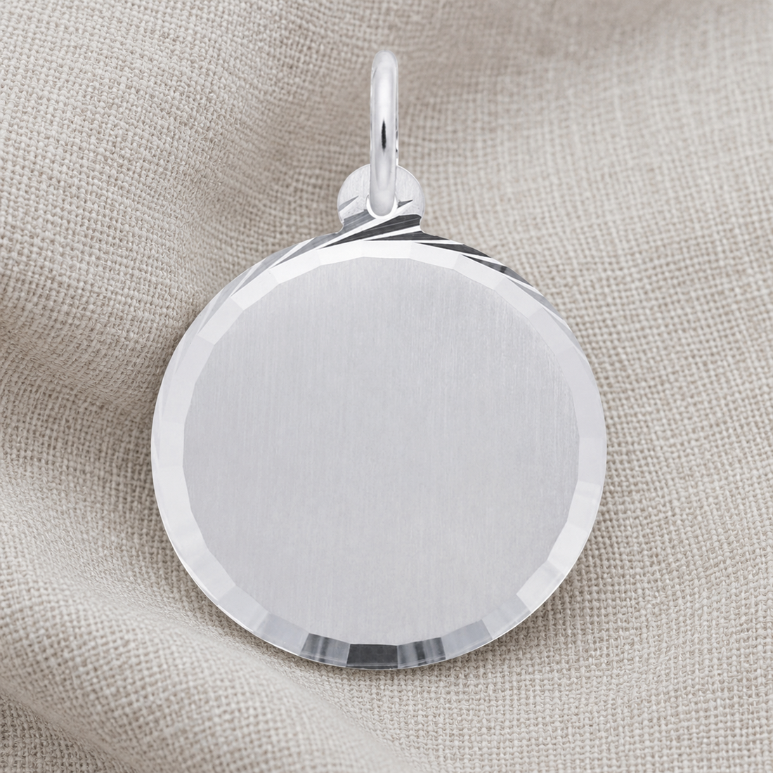 A Medium Diamond Faceted Disc - Sterling Silver Charm by Rembrandt Charms, featuring a smooth center and sparkling faceted edges, rests on light beige fabric. This engravable charm offers an elegant option for those looking to personalize their jewelry collection.