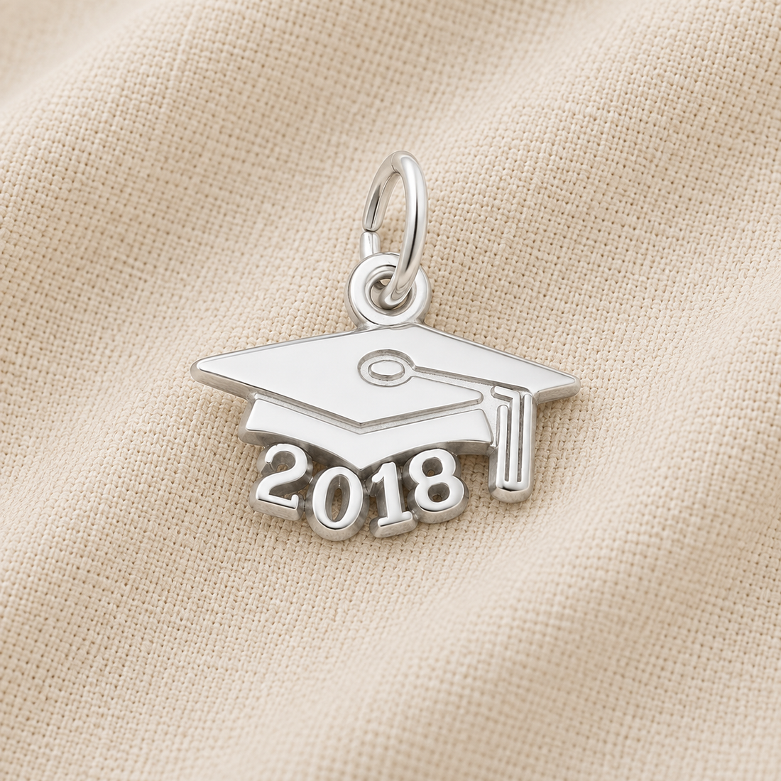 A Rembrandt Charms 2018 Grad Cap—Sterling Silver charm rests on a textured beige fabric background, making an ideal graduation keepsake. This classic piece features “2018” beneath the iconic cap design, beautifully commemorating the milestone.