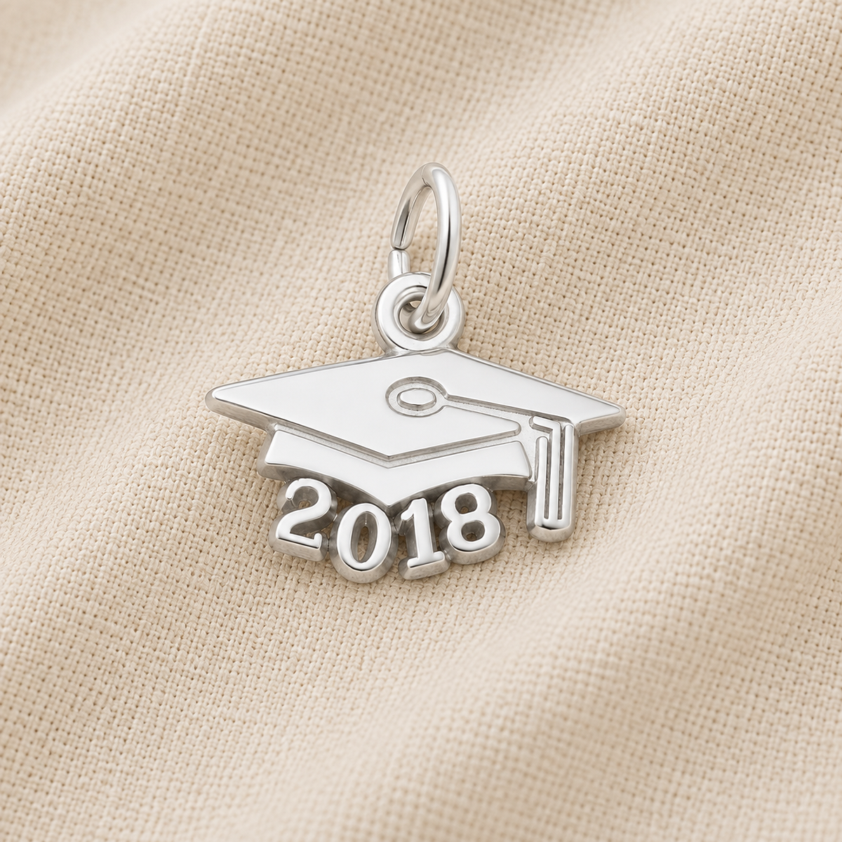 A Rembrandt Charms 2018 Grad Cap—Sterling Silver charm rests on a textured beige fabric background, making an ideal graduation keepsake. This classic piece features “2018” beneath the iconic cap design, beautifully commemorating the milestone.