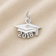 A Rembrandt Charms 2018 Grad Cap—Sterling Silver charm rests on a textured beige fabric background, making an ideal graduation keepsake. This classic piece features “2018” beneath the iconic cap design, beautifully commemorating the milestone.