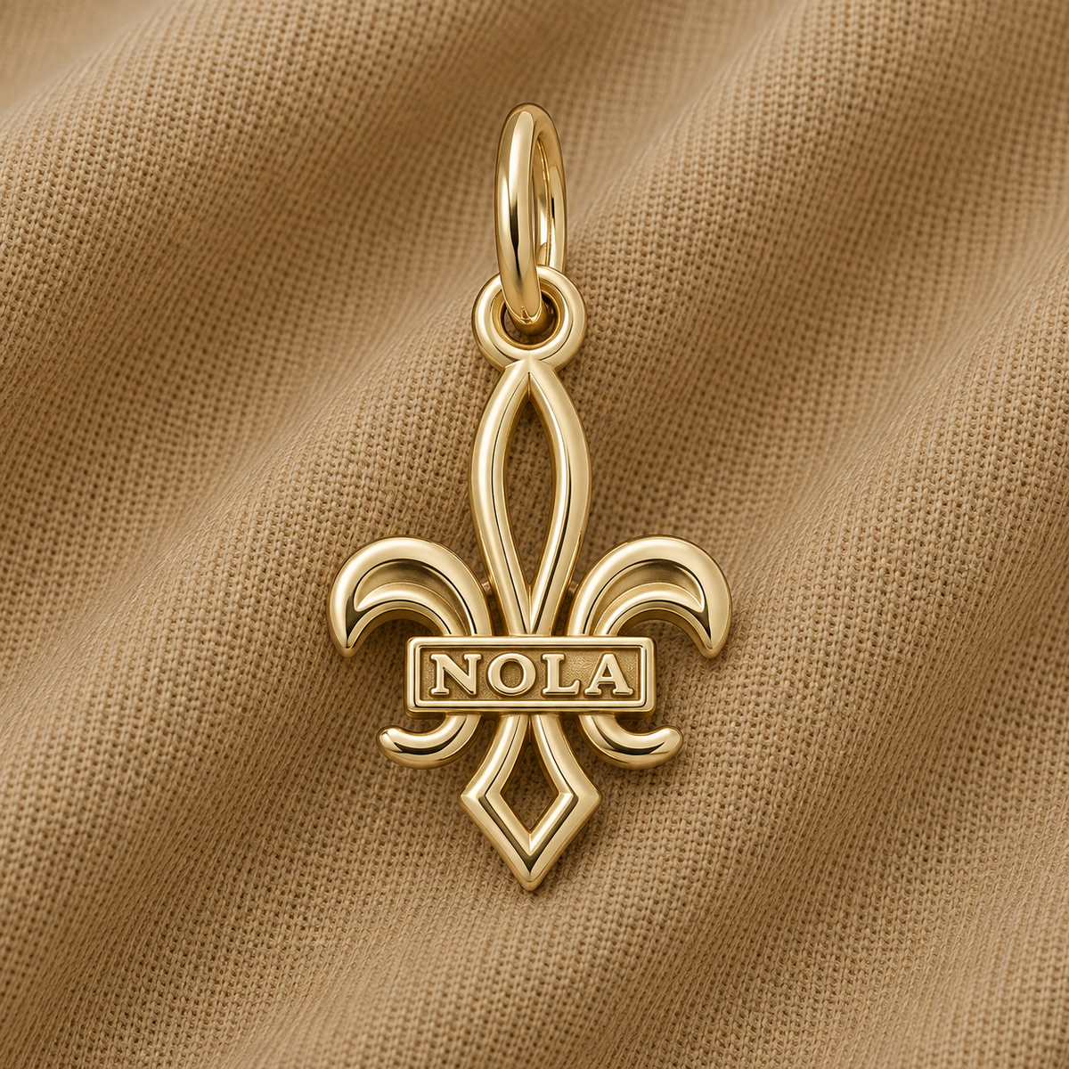 A gold-plated NOLA Fleur de Lis Charm by Rembrandt Charms, featuring “NOLA” across the center, displayed on a beige textured fabric—a classic New Orleans jewelry piece and a timeless Crescent City keepsake.