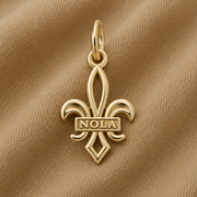 A gold-plated NOLA Fleur de Lis Charm by Rembrandt Charms, featuring “NOLA” across the center, displayed on a beige textured fabric—a classic New Orleans jewelry piece and a timeless Crescent City keepsake.