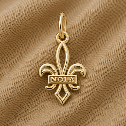 A gold-plated NOLA Fleur de Lis Charm by Rembrandt Charms, featuring “NOLA” across the center, displayed on a beige textured fabric—a classic New Orleans jewelry piece and a timeless Crescent City keepsake.