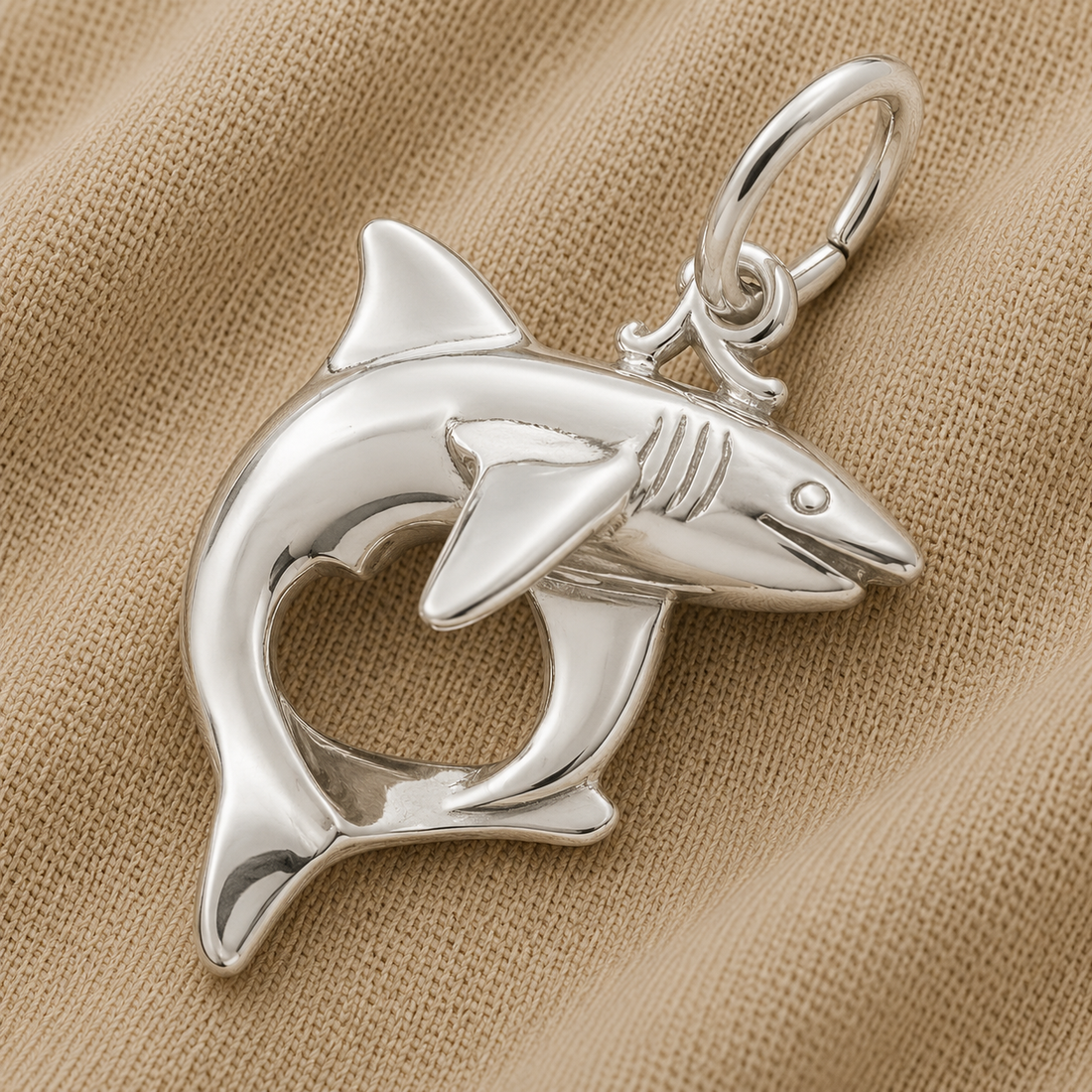The Rembrandt Charms Shark Charm in sterling silver features a striking shark-shaped pendant with a heart-shaped cutout in the body, beautifully displayed on beige textured fabric—an ideal three-dimensional piece of marine life jewelry for ocean lovers.