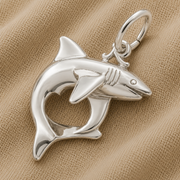 The Rembrandt Charms Shark Charm in sterling silver features a striking shark-shaped pendant with a heart-shaped cutout in the body, beautifully displayed on beige textured fabric—an ideal three-dimensional piece of marine life jewelry for ocean lovers.