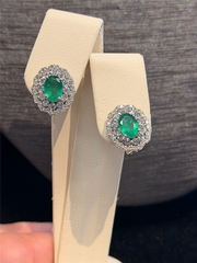 A pair of Ryan Gems Oval Emerald Earrings with Double Diamond Halo in 14k White Gold is displayed on a cream-colored stand against a dark background.