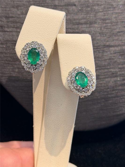 A pair of Ryan Gems Oval Emerald Earrings with Double Diamond Halo in 14k White Gold is displayed on a cream-colored stand against a dark background.