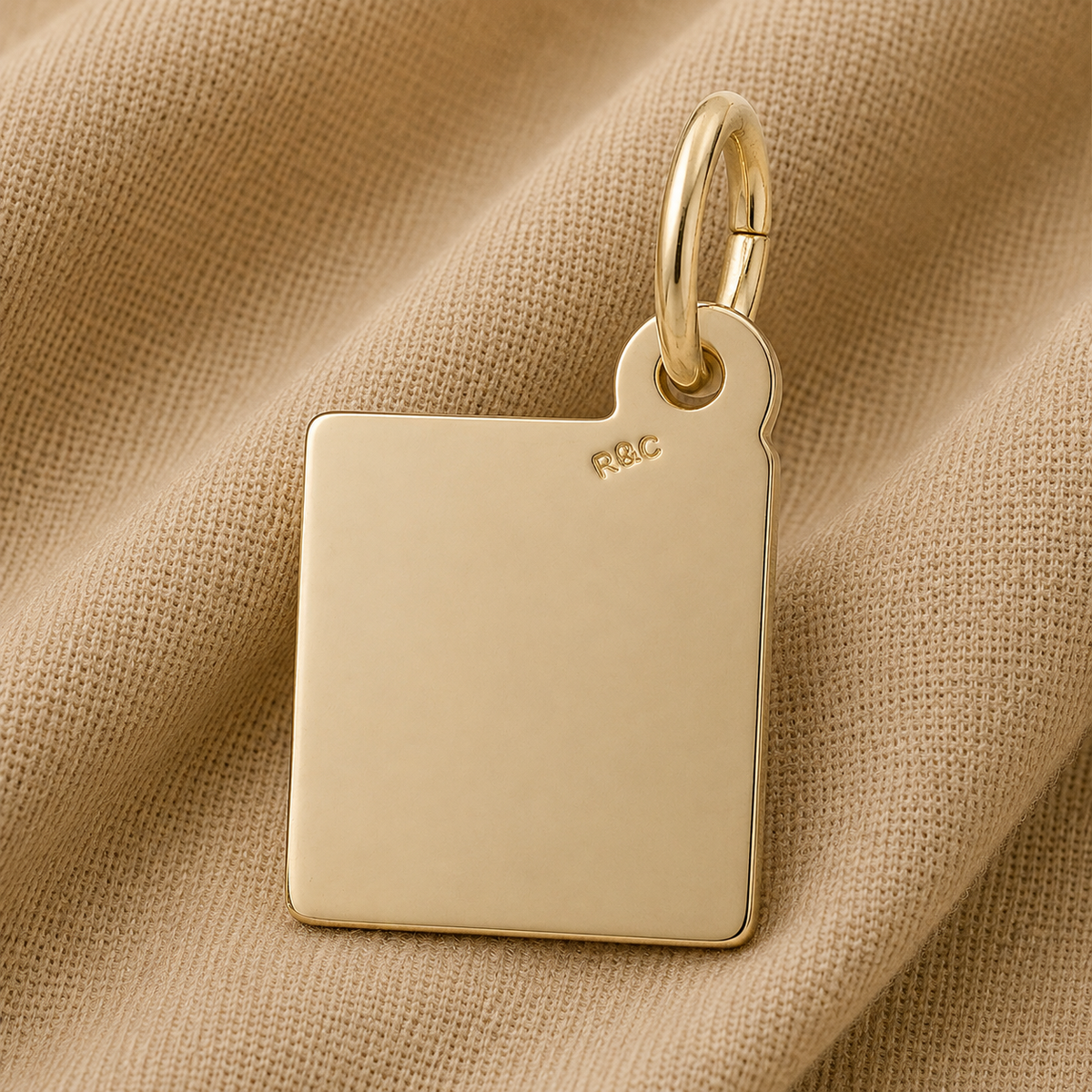A gold-plated, rectangular Bible Charm by Rembrandt Charms featuring a rounded corner and a delicate “R&C” engraving rests on beige textured fabric—an elegant piece of faith jewelry that brings subtle meaning to any collection.