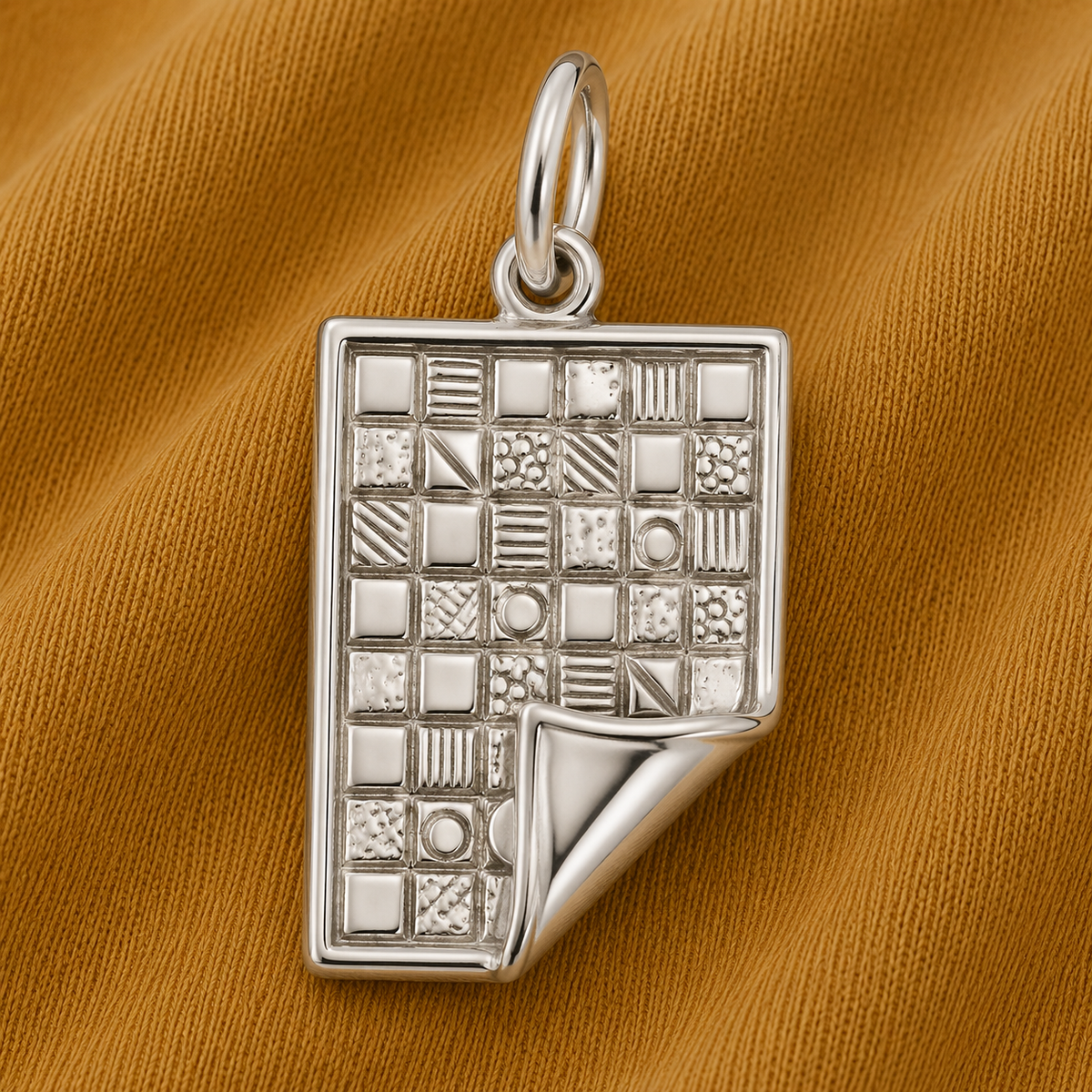 Showcasing a rectangular sterling silver pendant with a grid pattern and textured squares, plus a curled corner detail, the Quilt Charm by Rembrandt Charms is photographed on mustard yellow fabric—making it a distinctive gift for quilters and an ideal choice for anyone who loves handmade quilt jewelry.