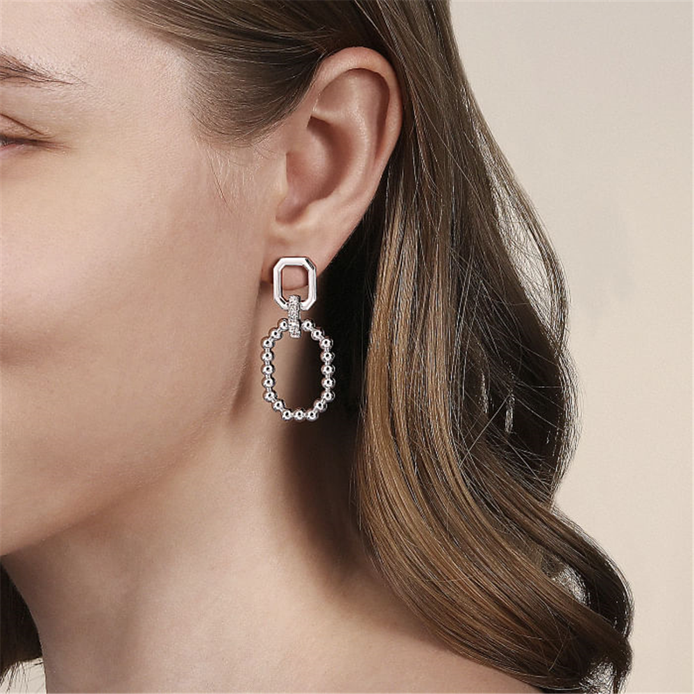 A close-up of a person wearing the sophisticated 925 Sterling Silver Octagon White Sapphire Bujukan Stud Drop Earring from Gabriel & Co. The earring features a distinctive small octagonal design connected to a larger oval-shaped loop adorned with delicate beads. The person's brown hair is partially visible, cascading in soft waves, against a neutral background.