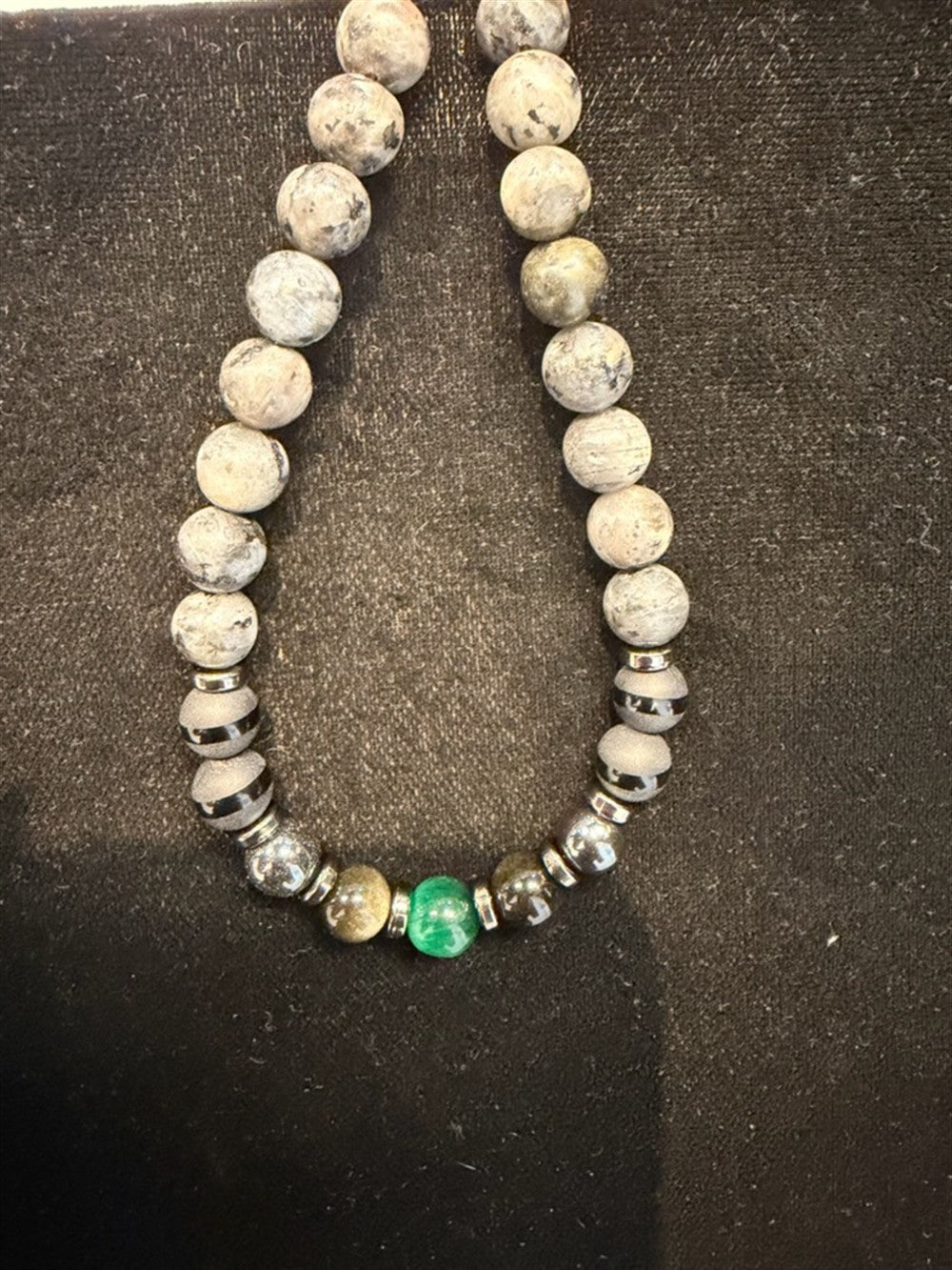 Redemption Jewelry’s “already sold - Liz” features round, speckled gray stones, metallic spacers, and a central green bead, displayed on a black surface.
