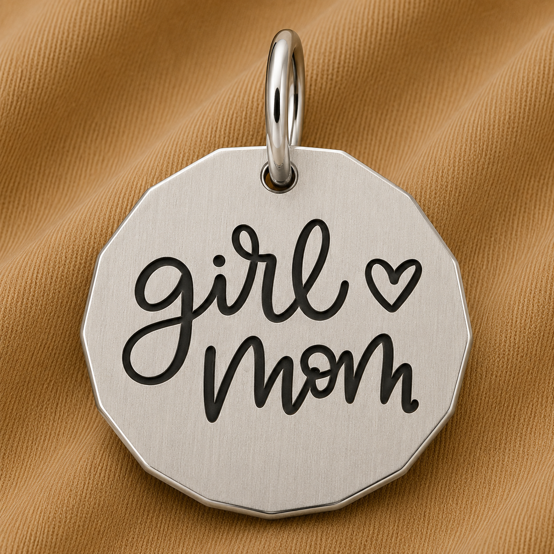 The Rembrandt Charms Girl Mom Charm in sterling silver, featuring “girl mom” and a heart symbol engraved on it, is displayed on a light brown textured fabric—this beautiful charm makes the perfect Mother’s Day gift and serves as a timeless piece of mother-daughter jewelry.