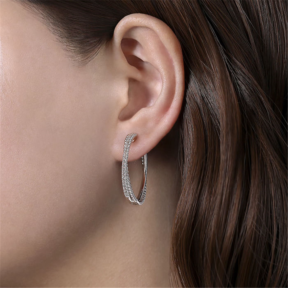 Close-up of the side of a person's head, showcasing their ear adorned with Gabriel & Co's 925 Sterling Silver White Sapphire Twisted Bujukan 30mm Classic Hoop. The person has brown hair draped behind the ear, highlighting the sparkling earring.