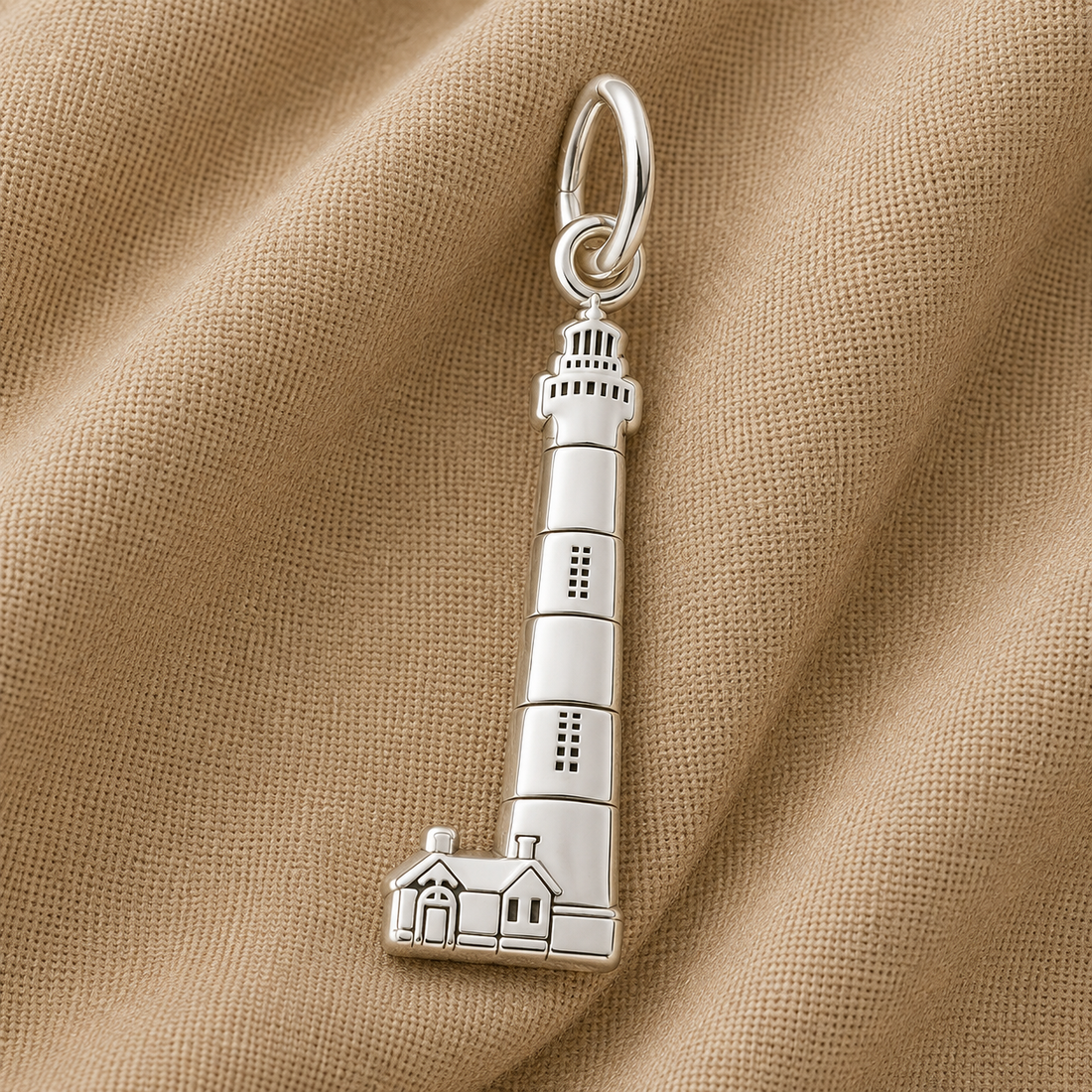 Showcasing exquisite Outer Banks jewelry, the Bodie Island Lighthouse Charm in sterling silver by Rembrandt Charms features a lighthouse-shaped design with two detailed houses at the base. Set against light brown textured fabric, this charm is a perfect coastal keepsake.
