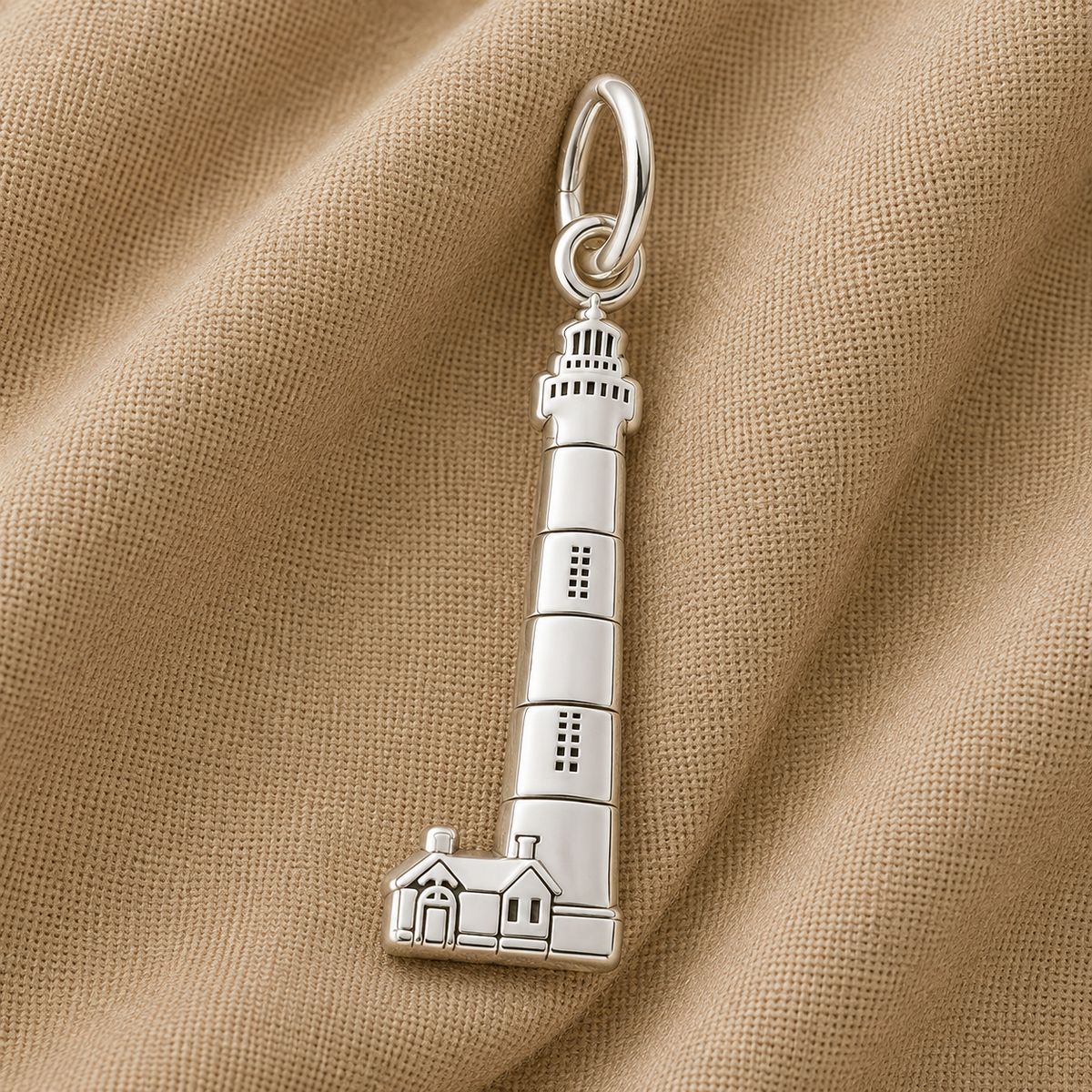 Showcasing exquisite Outer Banks jewelry, the Bodie Island Lighthouse Charm in sterling silver by Rembrandt Charms features a lighthouse-shaped design with two detailed houses at the base. Set against light brown textured fabric, this charm is a perfect coastal keepsake.