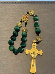 The SR005 Small Green Tiger Eye Rosary by Redemption Jewelry, featuring green beads, gold medallions, and a large gold crucifix with Jesus, is displayed on a gray surface.