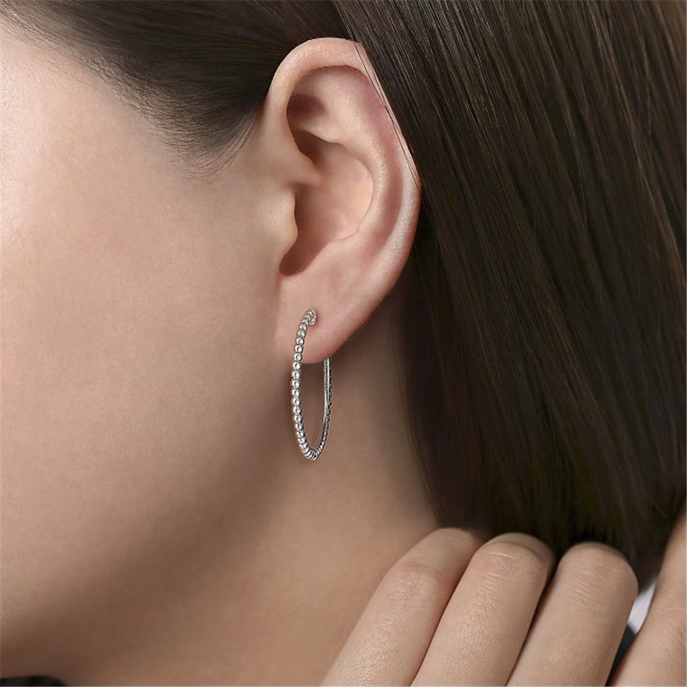 A close-up shows a person's ear adorned with Gabriel & Co's Sterling Silver Bujukan Classic Hoop Earrings | 30mm. The hoops are encrusted with small diamonds and crafted from 925 sterling silver. The person has straight, dark hair tucked behind the ear and touches their neck with their fingers.