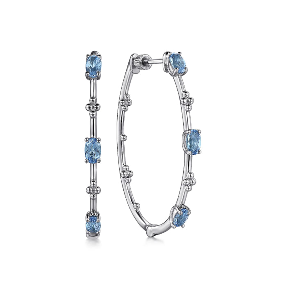 Gabriel & Co's 925 Sterling Silver Swiss Blue Topaz Bujukan Classic Hoop Earrings feature an elegant design with oval-shaped Swiss Blue Topaz gemstones. Adorned with small silver beads interspersed between the gemstones and set in a sleek silver frame, these sophisticated earrings are displayed upright against a plain white background.