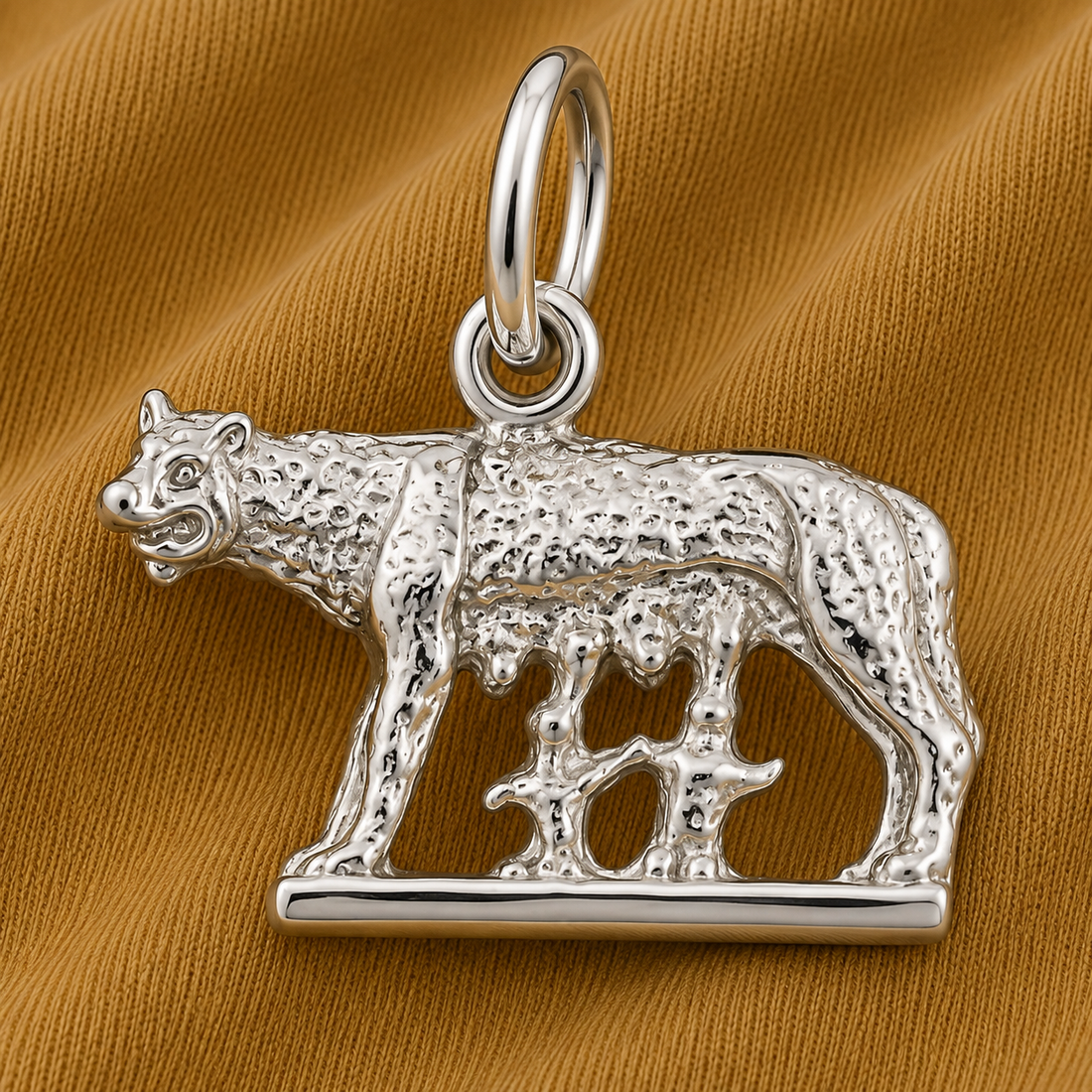 A sterling silver Romulus and Remus Charm by Rembrandt Charms, featuring a detailed she-wolf standing over two small figures, beautifully set against a textured golden fabric backdrop—an exquisite piece inspired by the legendary origins of ancient Rome.
