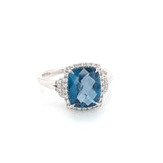 A striking piece from Bellarri, the "Large London Blue Topaz and Diamonds Ring" showcases a captivating 14K white gold band featuring a prominent cushion-cut London Blue Topaz gemstone at its center. The gemstone is elegantly surrounded by a halo of delicate white diamonds. For added sophistication, the band splits into two near the setting, all set against a plain white backdrop.