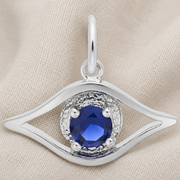 The Rembrandt Charms Evil Eye Charm- Sterling Silver, featuring an elegant eye-shaped design with a round blue gemstone at its center, is displayed on beige fabric.