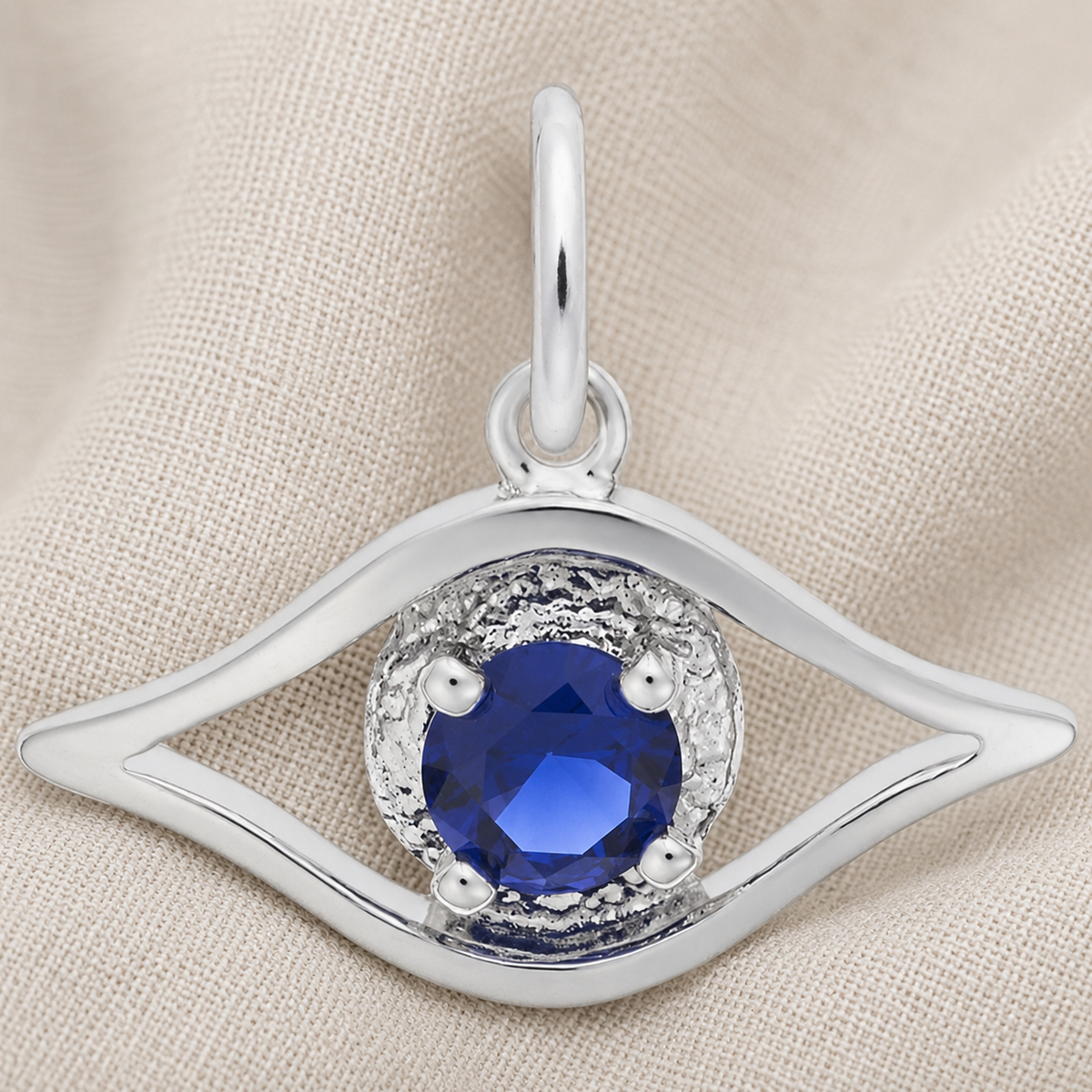 The Rembrandt Charms Evil Eye Charm- Sterling Silver, featuring an elegant eye-shaped design with a round blue gemstone at its center, is displayed on beige fabric.