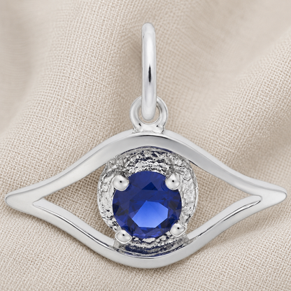 The Rembrandt Charms Evil Eye Charm- Sterling Silver, featuring an elegant eye-shaped design with a round blue gemstone at its center, is displayed on beige fabric.