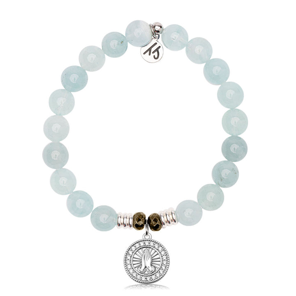 The T Jazelle Blue Aquamarine Gemstone Bracelet features light blue round beads, a sterling silver prayers charm with a CZ accent, and a small round metal tag engraved with a symbol.