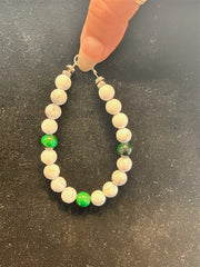 A hand holds Redemption Jewelry’s B001 Howlite & Green Tiger Eye Bracelet, featuring mostly white marbled howlite beads and four symmetrically arranged green tiger eye beads, resting on a dark surface.