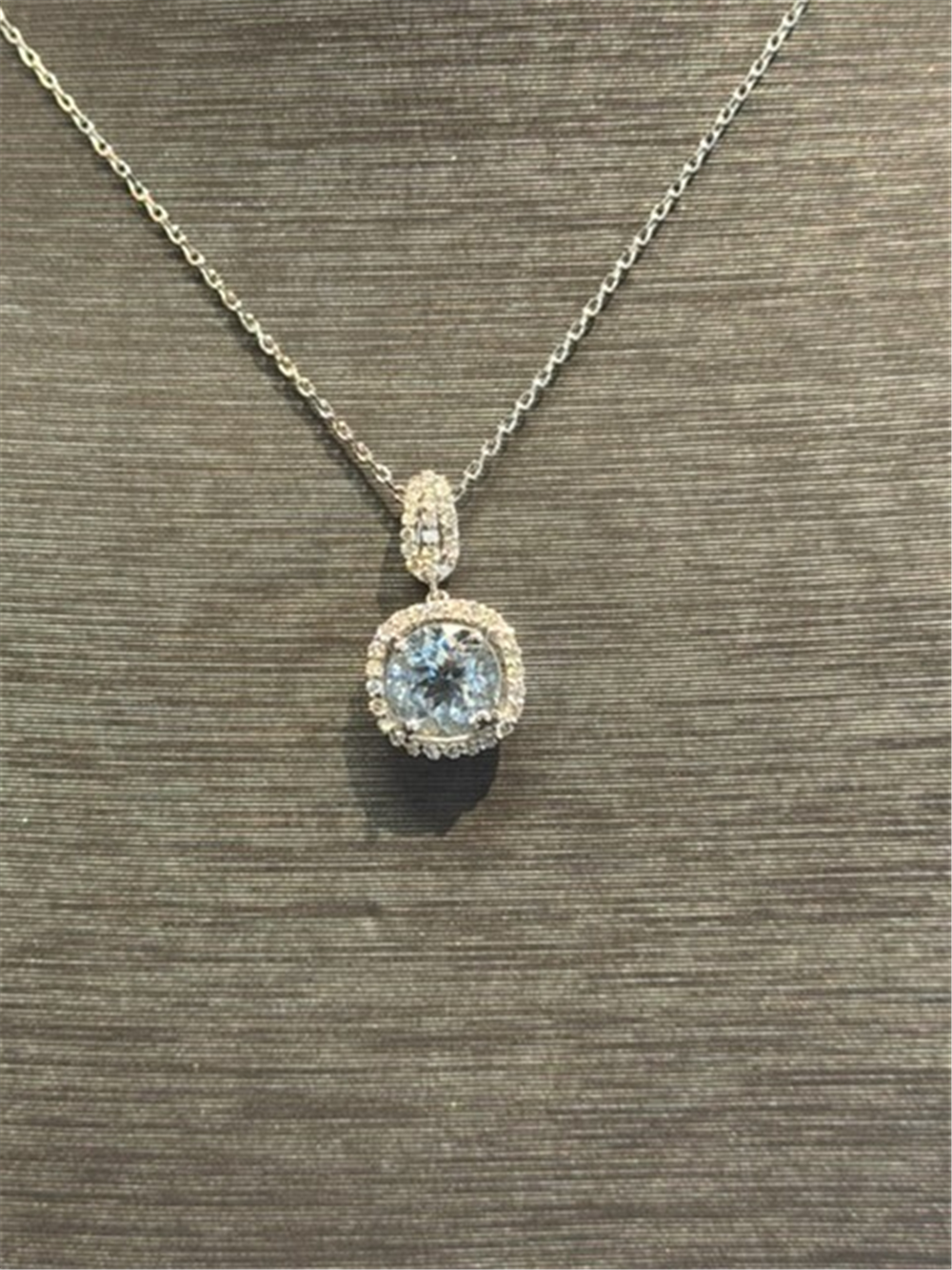 The Ryan Gems Aquamarine and Diamond Pendant in 14k white gold, featuring a round aquamarine surrounded by sparkling diamonds, is shown on a textured gray surface, highlighting its elegant beauty.