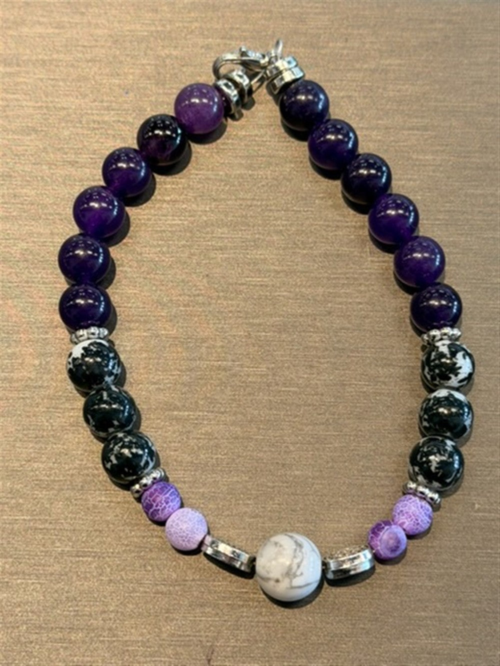 Redemption Jewelry’s B006 bracelet features Amethyst, Snowflake Obsidian, frosted Dragon Agate in purple, black, white, and a central marbled Howlite bead, shown on a brown textured surface.