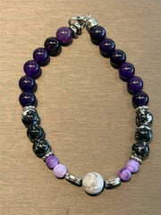 Redemption Jewelry’s B006 bracelet features Amethyst, Snowflake Obsidian, frosted Dragon Agate in purple, black, white, and a central marbled Howlite bead, shown on a brown textured surface.