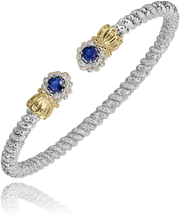 The Alwand Vahan Jewelry bracelet features an elegant open-ended design, crafted in a twisted rope style with a combination of 14k gold and sterling silver. It is adorned with two end caps, each showcasing a round blue sapphire encircled by smaller clear stones. Weighing in at 0.10 carats, the bracelet rests gracefully on the wrist, and its reflective surface highlights the intricate detailing.