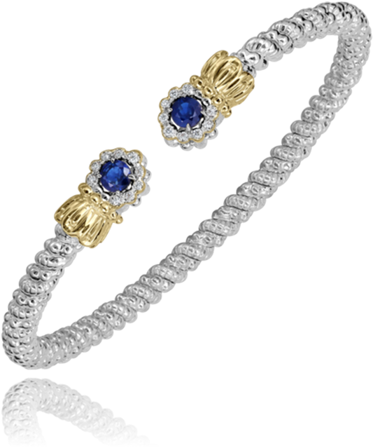 The Alwand Vahan Jewelry bracelet features an elegant open-ended design, crafted in a twisted rope style with a combination of 14k gold and sterling silver. It is adorned with two end caps, each showcasing a round blue sapphire encircled by smaller clear stones. Weighing in at 0.10 carats, the bracelet rests gracefully on the wrist, and its reflective surface highlights the intricate detailing.
