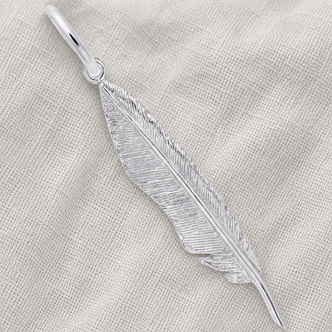 A Feather Charm - Sterling Silver by Rembrandt Charms rests on a textured, light beige fabric, highlighting the intricate etching and timeless elegance of this symbolic jewelry piece.