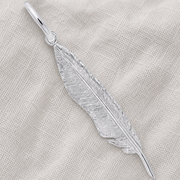 A Feather Charm - Sterling Silver by Rembrandt Charms rests on a textured, light beige fabric, highlighting the intricate etching and timeless elegance of this symbolic jewelry piece.