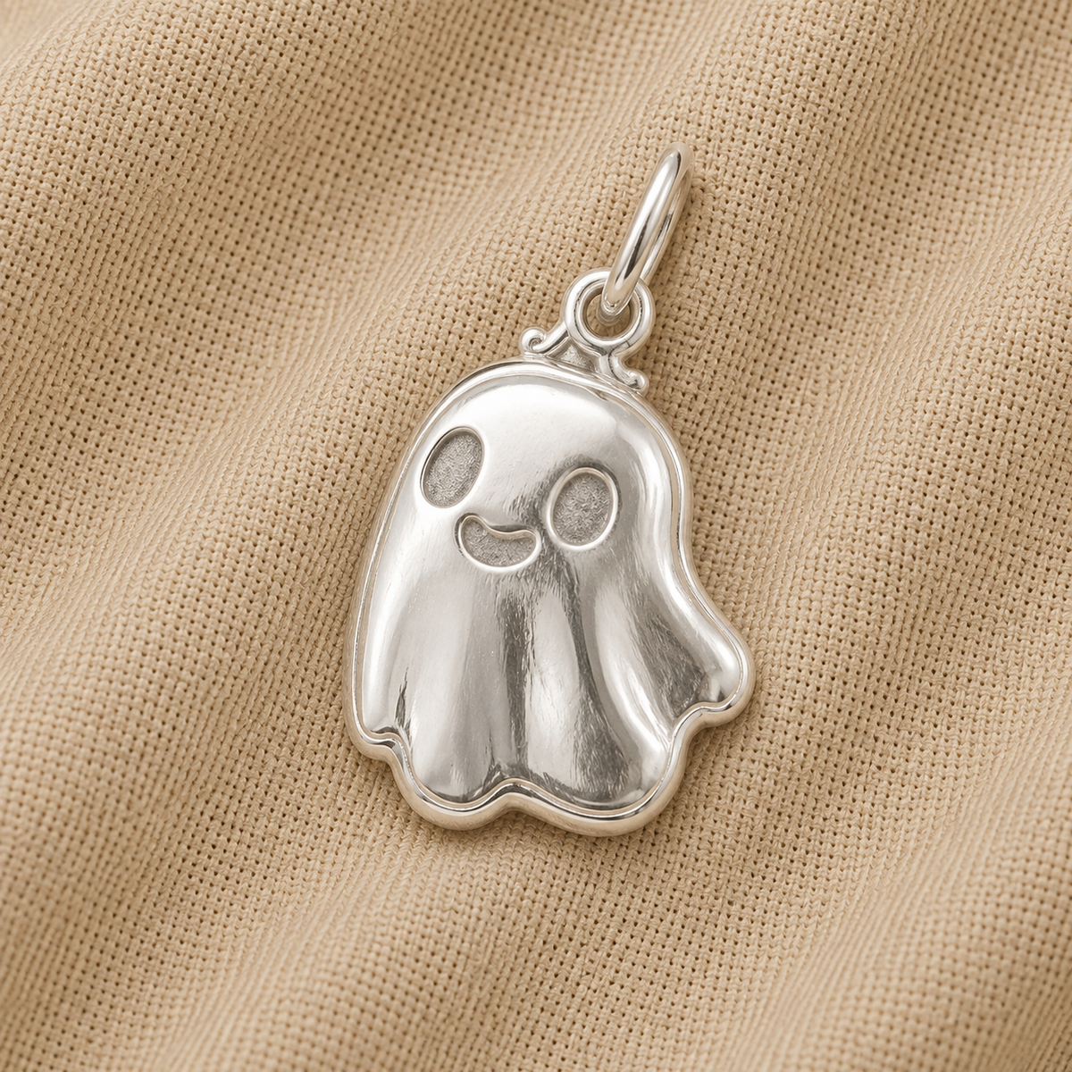 The Rembrandt Charms Cute Ghost Charm, crafted in sterling silver and featuring a smiling face, rests on beige textured fabric—an adorable piece of Halloween jewelry.