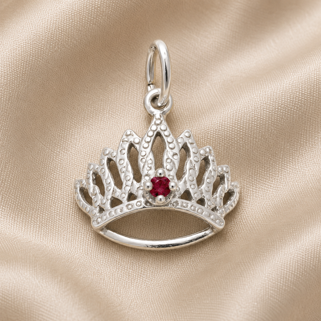 The Tiara With Birthstone Charm (January) by Rembrandt Charms, crafted in sterling silver and featuring a vivid red gemstone at its center, is elegantly displayed on a soft beige fabric background.