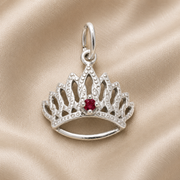 The Tiara With Birthstone Charm (January) by Rembrandt Charms, crafted in sterling silver and featuring a vivid red gemstone at its center, is elegantly displayed on a soft beige fabric background.
