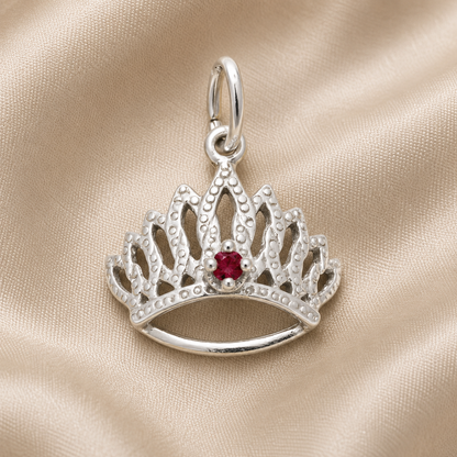 The Tiara With Birthstone Charm (January) by Rembrandt Charms, crafted in sterling silver and featuring a vivid red gemstone at its center, is elegantly displayed on a soft beige fabric background.