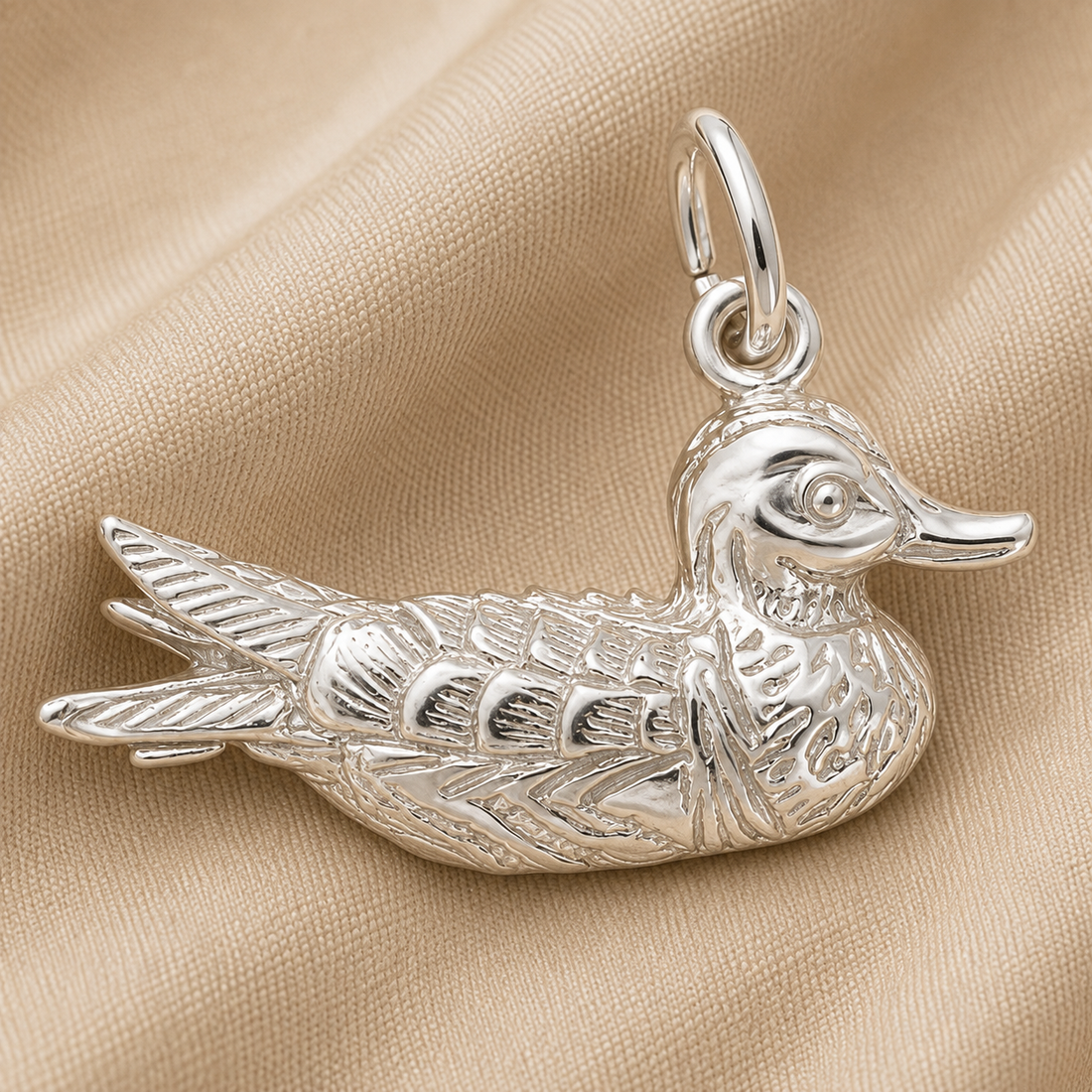 The Duck Charm—Sterling Silver by Rembrandt Charms features beautifully engraved details and is displayed on a soft beige fabric background, making it an adorable accessory ideal for animal lovers and charm collectors alike.