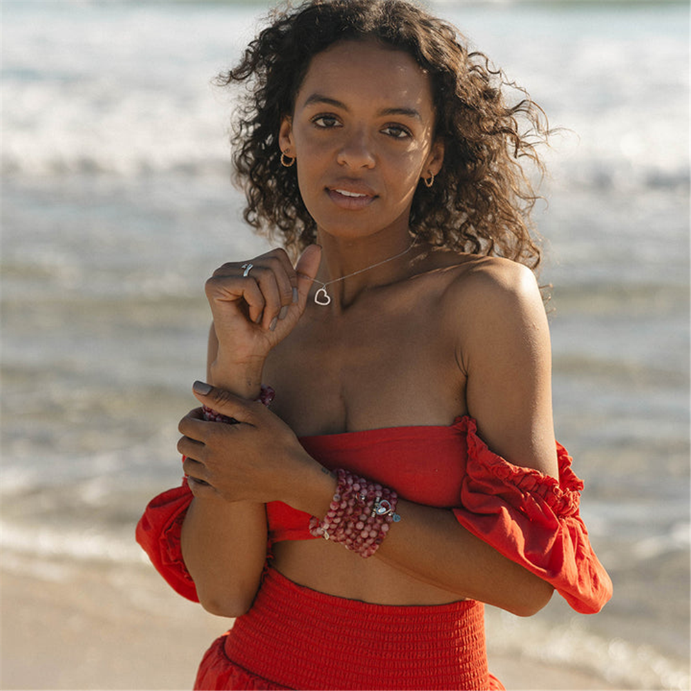 On a sunlit beach, a woman in a red off-shoulder top and skirt holds the T Jazelle Beautiful Soul Gold Charm Necklace, with ocean waves shimmering in the background, embodying reflection and grace.