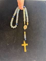 A hand holds the LR005 Long Crystal & Blue Tiger Eye Rosary by Redemption Jewelry, featuring iridescent beads, a gold-colored crucifix, and a small medallion against a black background.