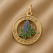 The Rembrandt Charms Texas Bluebonnets Charm - Gold Plated features a gold pendant on fabric, showcasing a bluebonnet flower at its center with “TEXAS BLUEBONNETS” engraved around the edge in capital letters. This elegant piece beautifully captures the natural beauty and state pride of Texas, making it a meaningful addition to any jewelry collection.