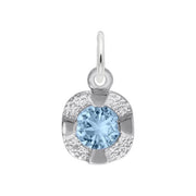 A close-up image of the Petite Birthstone - December Charm by Rembrandt Charms, crafted in sterling silver. This pendant features a round, blue birthstone at its center, surrounded by a textured silver design set in a slight cushion shape. It includes a loop at the top for easy attachment to a chain or necklace, making it an ideal gift for someone with a December birthday.