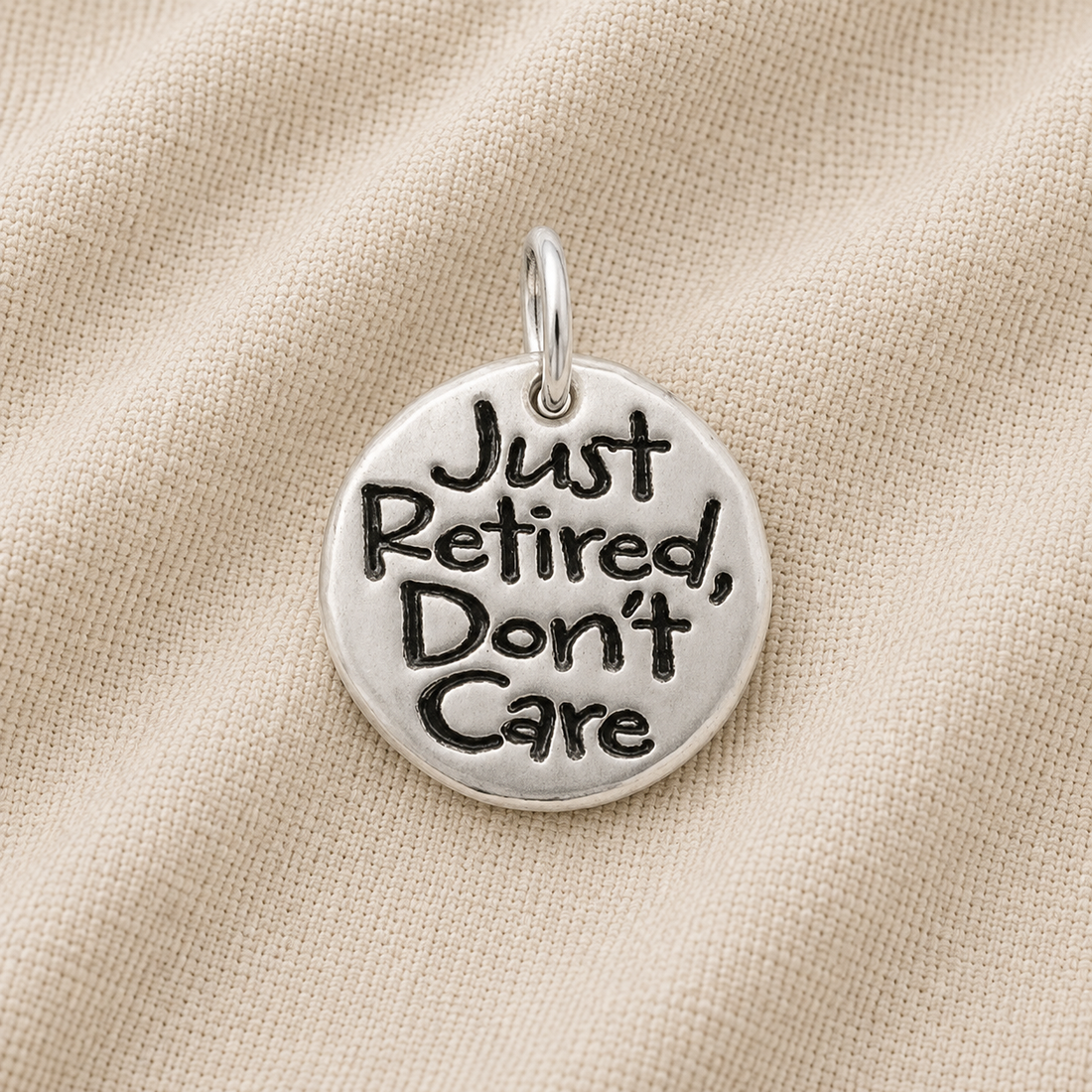 The Rembrandt Charms Just Retired disc sterling silver charm, featuring the inscription “Just Retired, Don’t Care,” is displayed against a beige fabric background—an ideal and unique addition to any charm bracelet or a thoughtful retirement gift.