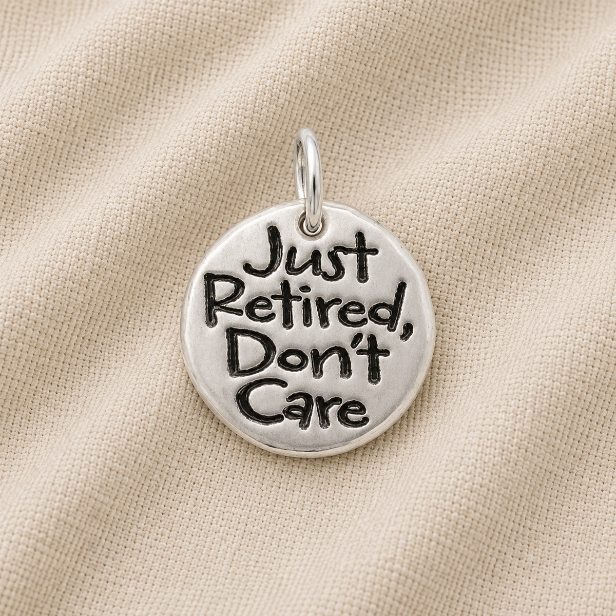 The Rembrandt Charms Just Retired disc sterling silver charm, featuring the inscription “Just Retired, Don’t Care,” is displayed against a beige fabric background—an ideal and unique addition to any charm bracelet or a thoughtful retirement gift.